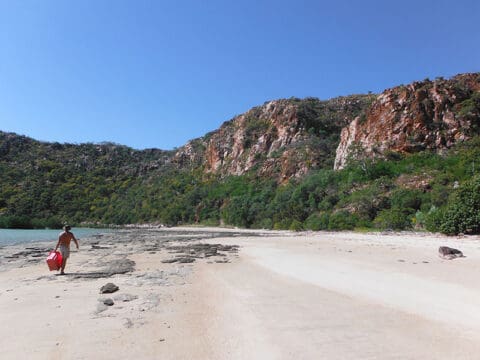 A person carrying a red cooler walks along a deserted beach with rocky terrain and shrub-covered cliffs under a clear blue sky, dreaming of One Tide Charters: private boat rentals for the ultimate escape.