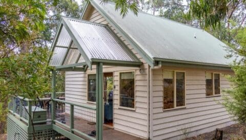 A wooden cabin with a green roof is surrounded by trees. It has a small porch, multiple windows, and a staircase leading up to the entrance. This quaint Bush House Cottage is nestled in Lorne’s forested area, making it an ideal eco retreat.