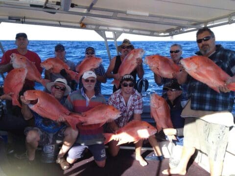 A group of ten men aboard a Seaforce boat are holding large, red fish, posing against the stunning backdrop of Coral Bay's ocean.