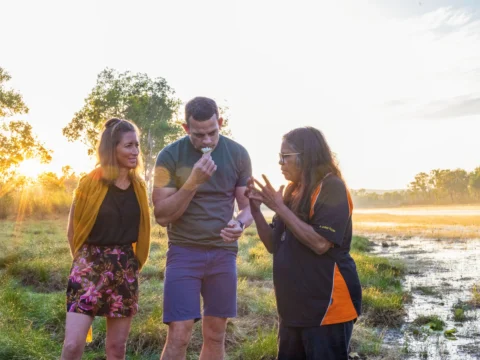 Three people are outdoors near a wetland area during sunrise or sunset. One person is holding and smelling a plant, while another gestures as if explaining something about sustainable ecotourism.