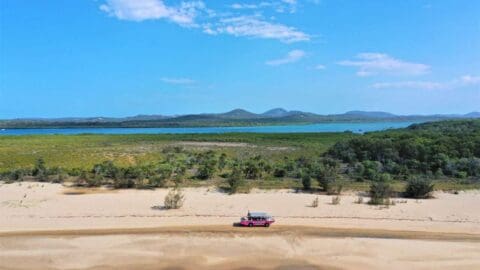 A sustainable pink vehicle travels on a sandy road with a lush landscape, body of water, and mountains in the background under a clear blue sky.