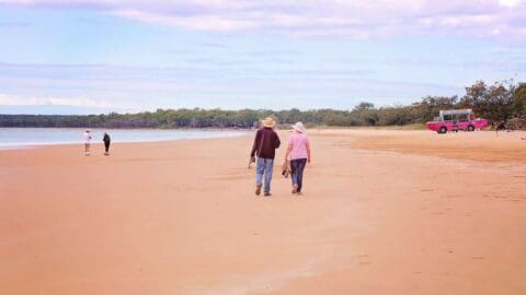 A couple walks hand-in-hand along a wide, empty beach, enjoying the serene beauty of nature while a few other people and a pink vehicle are visible in the background.