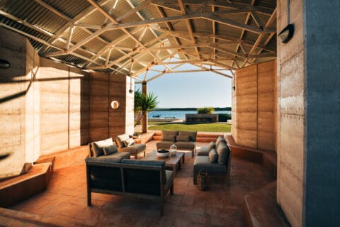 An open-air lounge area with cushioned seating under a sustainable, modern, pitched roof structure, overlooking a waterfront view.