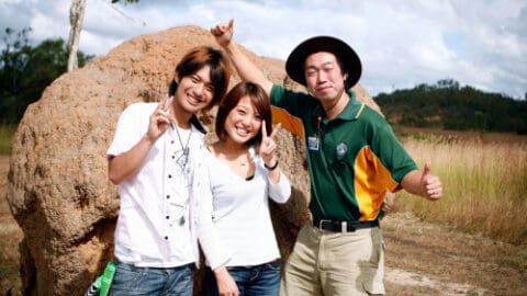 Three people posing and smiling in front of a large rock in an outdoor setting, with two of them making peace signs and one giving a thumbs-up. Courtesy of Doki Doki Tours, they are standing on a grassy area with trees in the background, enjoying their adventure.
