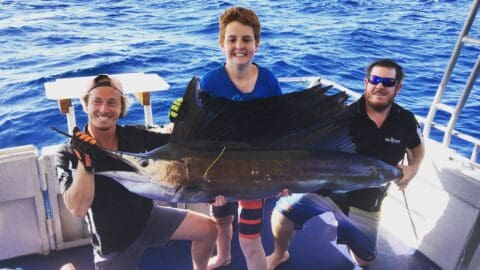 Three people on a boat proudly hold a large sailfish they caught in the waters of Coral Bay. The ocean shimmers brilliantly in the background.