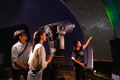 Three people at the World Nature Centre's observatory look at the night sky; one points with a green laser while another operates a telescope.