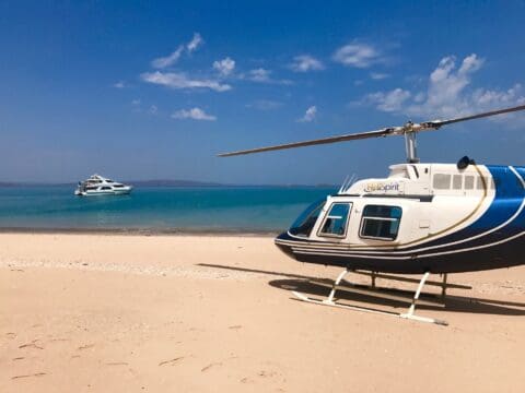 A chartered white helicopter is parked on a sandy beach near a calm sea, with a luxury yacht anchored in the distance under a clear blue sky.