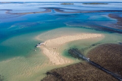 Aerial view of a small sandbar in turquoise waters with a few charter boats anchored and people walking on the sand and swimming. Islands and water channels are visible in the distance, showcasing nature's diversity.