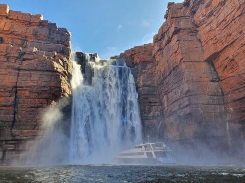 A boat sails near a large cascading waterfall surrounded by towering red rock cliffs under a clear blue sky, offering a glimpse of the breathtaking scenery enjoyed during the Lady M luxury cruises.