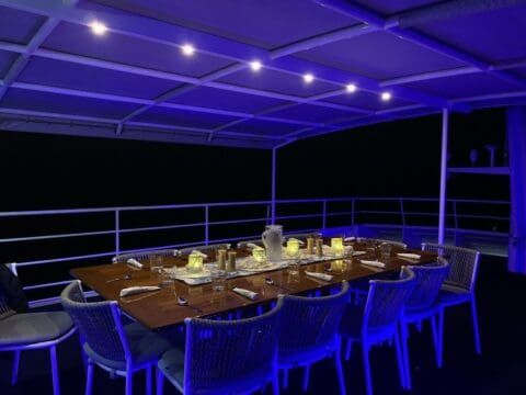 A dining table on a covered patio is set with place settings and candles, illuminated by blue lighting in a darkened setting, creating a perfect ambiance for a company gathering.