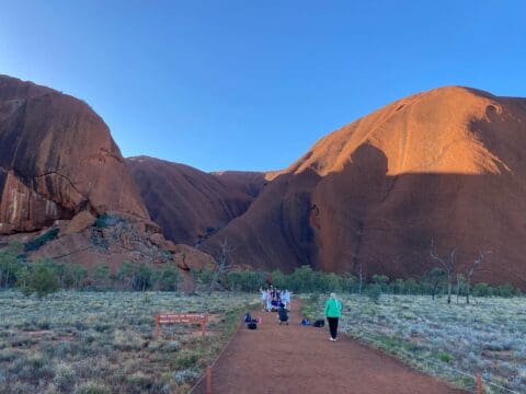 People walk towards a group gathered at the base of a large, rocky outcrop under a clear blue sky, reminiscent of an Earth Sanctuary.