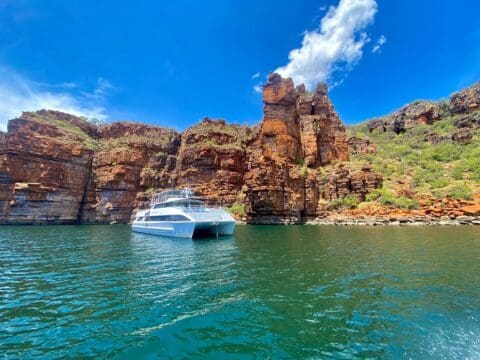 A white yacht, available for charter, is anchored in clear blue water surrounded by large, rugged canyon walls under a bright blue sky.