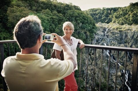 A person takes a photo of another person smiling and posing on a railing with a scenic view of a waterfall and forest in the background, capturing a memorable moment with Doki Doki Tours.