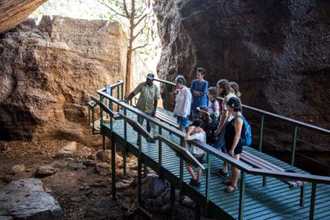 A group of people standing on a wooden platform inside a cave, listening attentively to a guide from Ayal Aboriginal Tours as they point at rock formations in Kakadu.