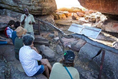 A tour guide from Ayal Aboriginal Tours explains to a group of people at an outdoor historical site in Kakadu, showcasing impressive rock formations and detailed informational signs.