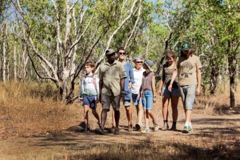 A group of six people, including a guide from Ayal Aboriginal Tours, walking along a dirt path in a wooded area with dry grass and tall trees on a sunny day in Kakadu.