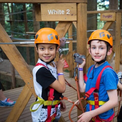 Two children wearing orange helmets and harnesses stand by a wooden structure labeled "White 2," preparing for an adventure on the TreeTops ropes course. Both are holding carabiners attached to safety cables, ready to explore the Central Coast from above.