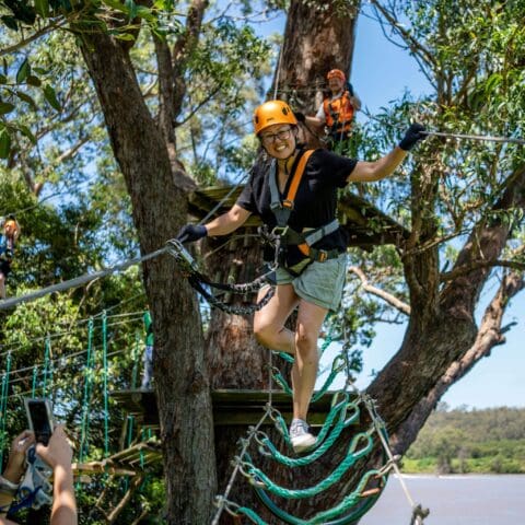 A person wearing a helmet and harness is crossing a rope bridge in the Central Coast's TreeTops Adventure park. They are smiling while holding onto the ropes, with others visible on platforms in the background.