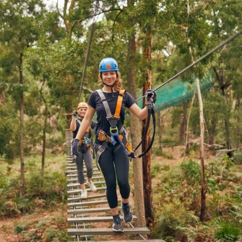 Two individuals wearing helmets and harnesses navigate a rope bridge in a forested area, engaged in an outdoor adventure activity at TreeTops Adventure on the Central Coast.