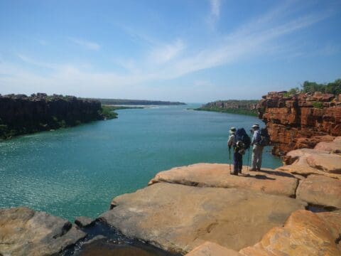 Three people in hiking attire, possibly part of Willis's Walkabouts, stand on the edge of a rocky cliff overlooking a wide river and scenic landscape under a clear blue sky.