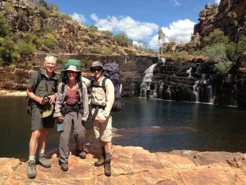 Three hikers with backpacks stand on rocky terrain in front of a waterfall and a clear pond, surrounded by a mountainous landscape under a partly cloudy sky, epitomizing the essence of Willis's Walkabouts' adventure travel.