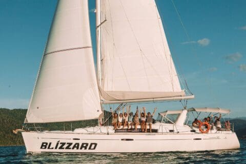 A group of people sit on the side of a white sailboat named "Blizzard" with sails raised, enjoying a day of sailing on the serene waters of the Whitsundays, with a hilly coastline in the background.