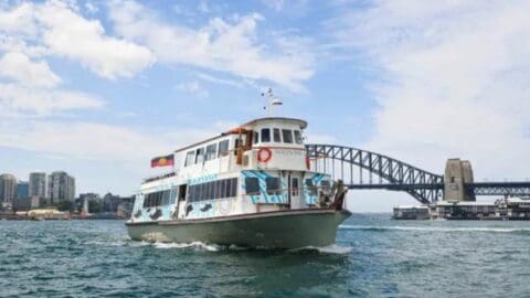 A ferry sails in the water near the Sydney Harbour Bridge on a clear day with a few scattered clouds, gliding like a tribal warrior through the serene landscape.