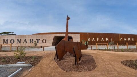 The entrance of Monarto Safari Park features modern architecture and metal animal sculptures under a clear sky, showcasing its commitment to ecotourism.