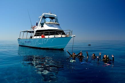 A boat is anchored in clear blue water with five people in wetsuits floating and waving near it, enjoying the beauty of nature.