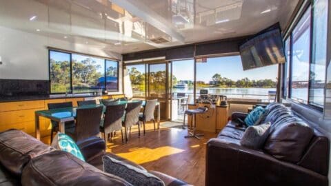 Bright modern houseboat interior with large windows, a dining area, a leather couch, and views of the water and trees outside. Perfect for an ecotourism adventure immersed in nature.