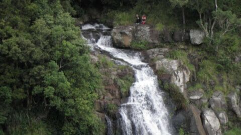 A waterfall cascades over rocky terrain surrounded by dense greenery. Two people stand on a rocky outcrop near the top of the waterfall, immersed in nature.