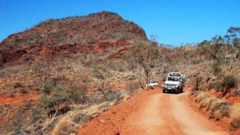 Two off-road vehicles tour a rugged, red rocky landscape with sparse vegetation, navigating a dirt track under a clear blue sky. In the background, a large hill stands majestically amid nature's serene beauty.