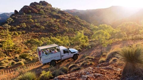 A white tour truck with passengers drives through a rugged, mountainous landscape with scattered greenery and the sun setting in the background, showcasing an authentic ecotourism experience.