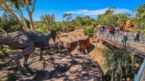 A group of people admire dinosaur sculptures from a bridge in a sunlit outdoor park, surrounded by trees and rocky terrain, embracing nature and the local ecotourism.