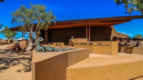 Outdoor exhibit featuring a bronze lizard sculpture on brown concrete platforms with a modernist building and trees under a clear blue sky, offering a touch of nature in an urban setting.