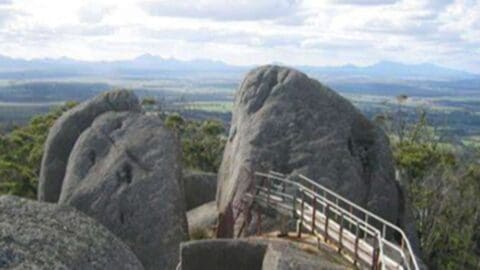 A metal handrail pathway leads through large, rounded rocks on a hill with a vast landscape of fields and distant mountains visible in the background under a cloudy sky, offering an ideal route for sustainable ecotourism.