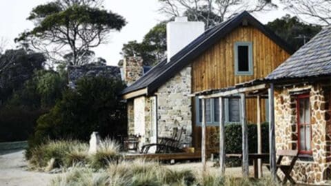 A rustic countryside house with stone and wood exterior, surrounded by tall grass and trees, offers charming accommodation. Featuring a small porch with wooden chairs, this ecotourism retreat is perfect for a relaxing escape.