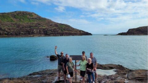 A group of six people stands on rocky terrain near the ocean with hills in the background, showcasing pristine nature. One person waves at the camera under a partly cloudy sky.