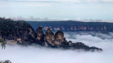 The Three Sisters, three rocky peaks, rise from a fog-covered valley with forested cliffs in the background, offering a stunning backdrop for your nature tour.