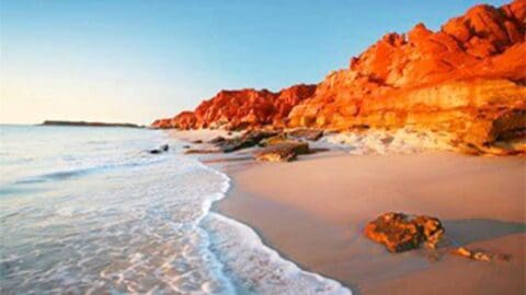 A tranquil beach scene with gentle waves washing onto a sandy shore, bordered by rugged red cliffs under a bright sky, invites you to explore the beauty of sustainable nature.