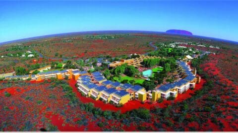 Aerial view of a resort complex with solar panels in a desert landscape, surrounding buildings, lush green spaces, and a distant large rock formation under a clear blue sky. This stunning tour destination offers exceptional accommodation nestled in the serenity of nature.