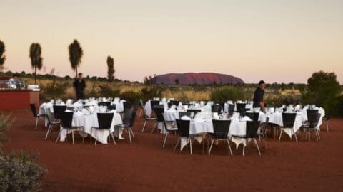 Ecotourism outdoor dining setup with multiple round tables covered in white tablecloths and black chairs arranged on red dirt ground, offering a distant view of a large rock formation at sunset.