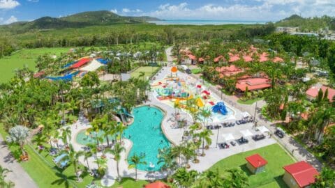 Aerial view of a resort featuring a large outdoor pool, surrounding lounge chairs, colorful water slides, and lush landscaping with trees and hills in the background under a sunny sky. This accommodation offers an inviting retreat for relaxation and ecotourism exploration.