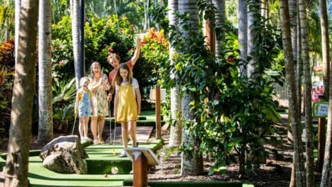 A group of people, including children, are enjoying a mini-golf tour in a lush, tropical setting with tall trees and vibrant greenery. One person is holding a golf club, and a golf ball is near a hole—an ideal spot for ecotourism enthusiasts.