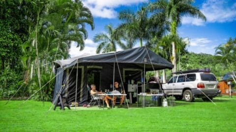 Two people sit under a large outdoor tent equipped with camping gear, next to an SUV, in a lush, nature-filled area with palm trees and a clear blue sky.