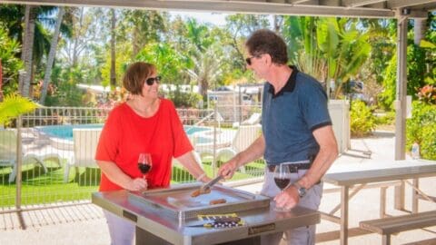 A man and woman enjoy a barbecue outside on a sunny day, holding glasses of wine and talking. Palm trees and a pool are visible in the background, enhancing the relaxed vibe. It’s the perfect setting for discussing their recent ecotourism tour adventures.