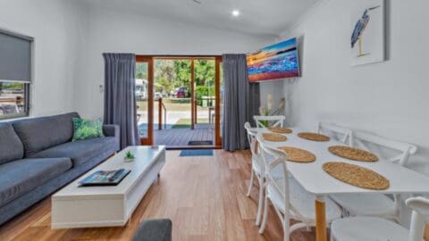 A bright living and dining area with a gray sofa, white coffee table, and a white dining table set with wicker placemats. A wall-mounted TV and a sliding glass door lead to a deck that offers tour-worthy views of nature.