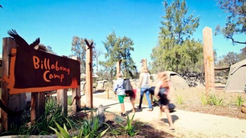 A wooden sign for Billabong Camp next to a path to a camp site that a family walks on it towards the camp.