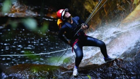 A person wearing a helmet and harness rappels down a waterfall, surrounded by flowing water and wet rocks, showcasing the thrill of ecotourism.