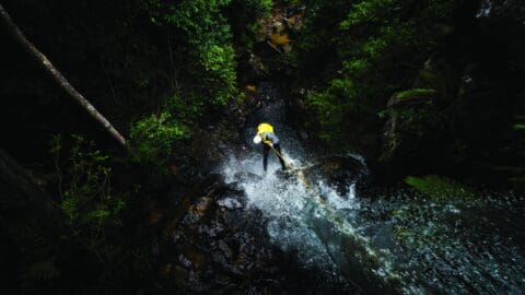 A person is rappelling down a waterfall surrounded by lush greenery, wearing a yellow helmet and safety gear. This thrilling ecotourism adventure offers an unforgettable experience as the water cascades down the rocks beneath them.